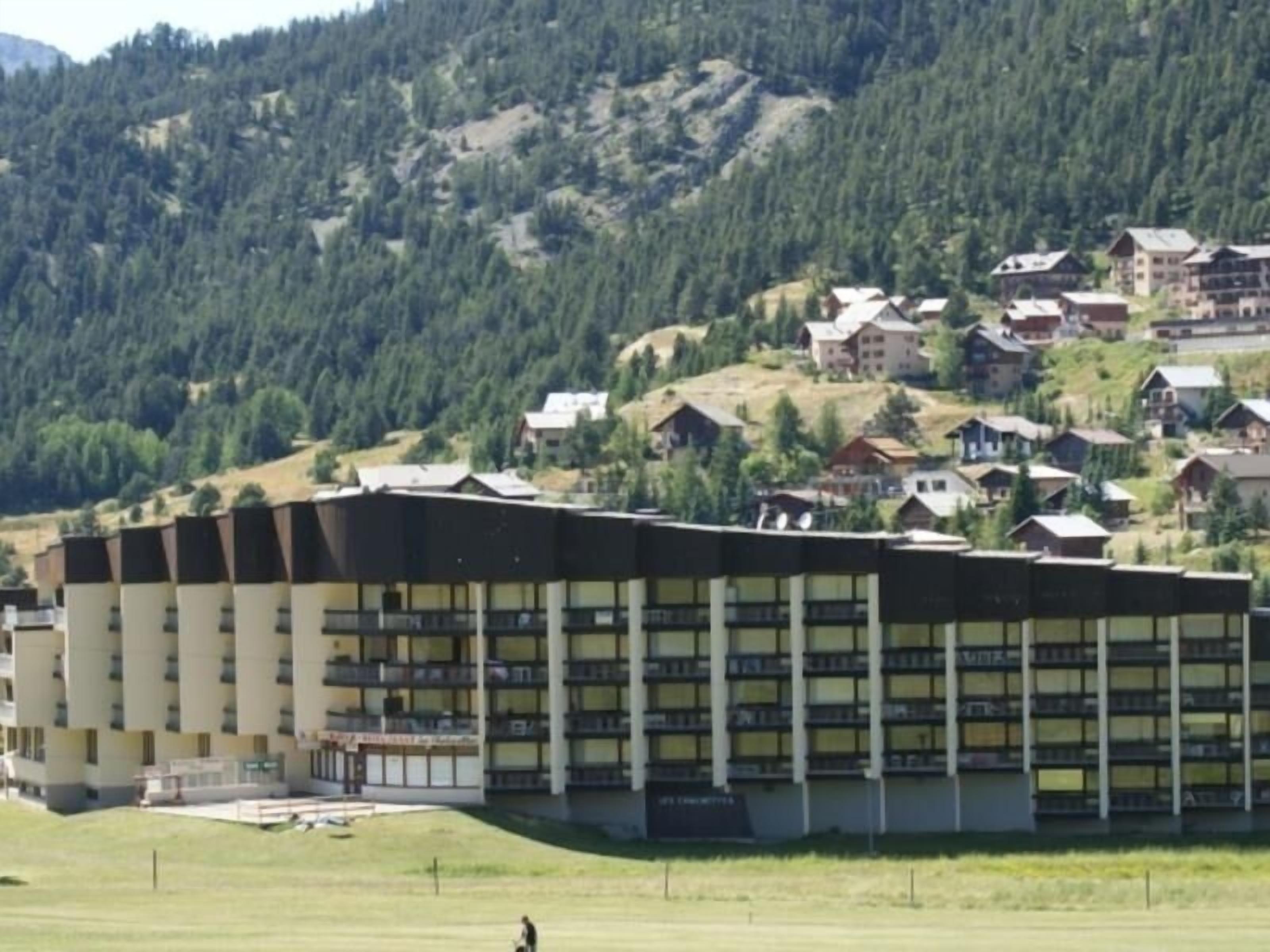 Un hôtel moderne doté d’un toit plat et de nombreux balcons, entouré d’un paysage verdoyant luxuriant et bordé, à l’arrière-plan, par une colline boisée.