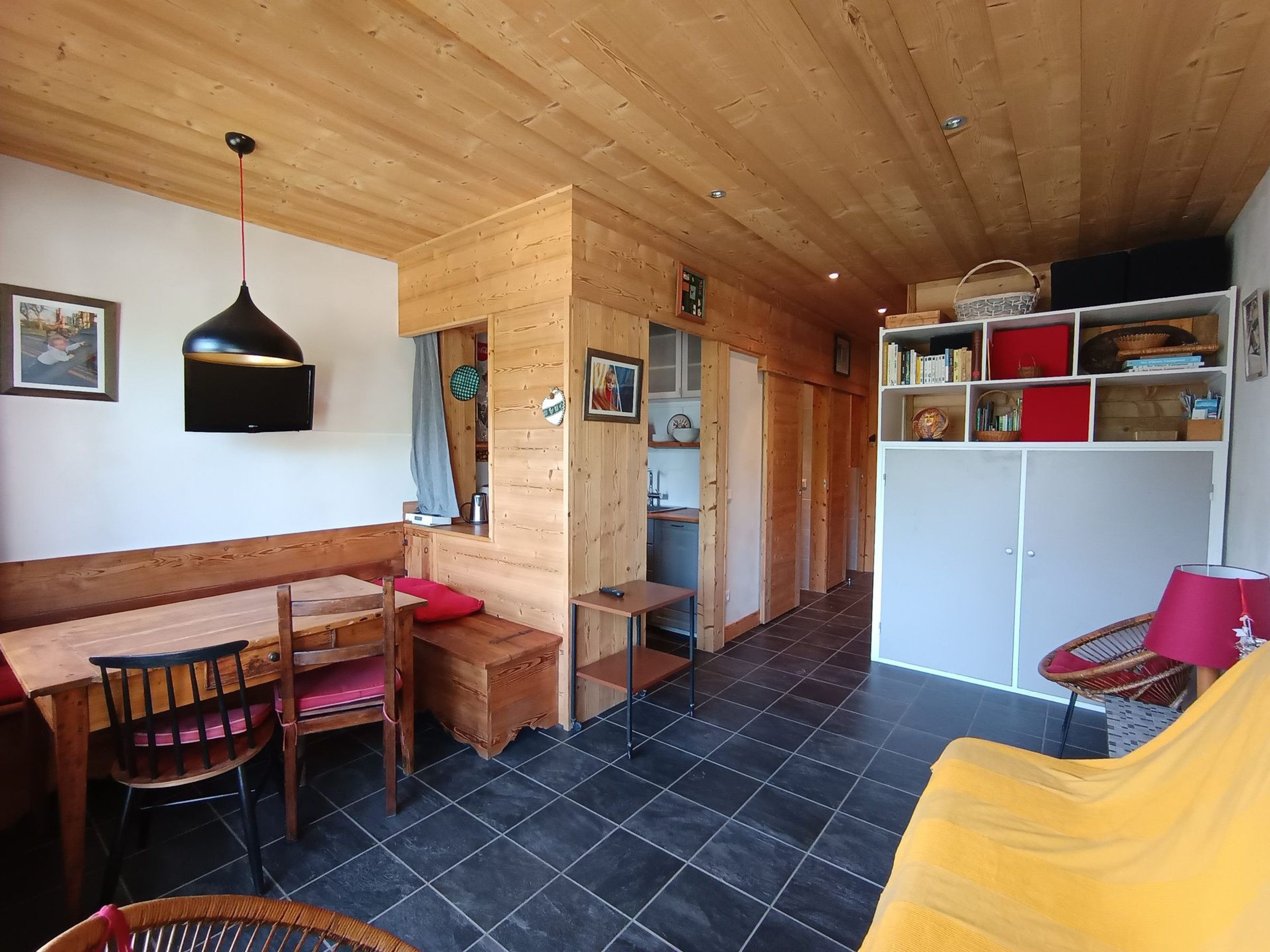 Una habitación acogedora con techo de madera, un comedor con banco y sillas, una estantería y una pequeña cocina.