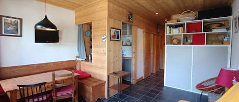 Una habitación acogedora con techo de madera, un comedor con banco y sillas, una estantería y una pequeña cocina.