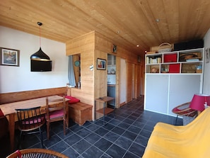 A cozy room with a wooden ceiling, a dining area with a bench and chairs, a bookshelf, and a small kitchen area.