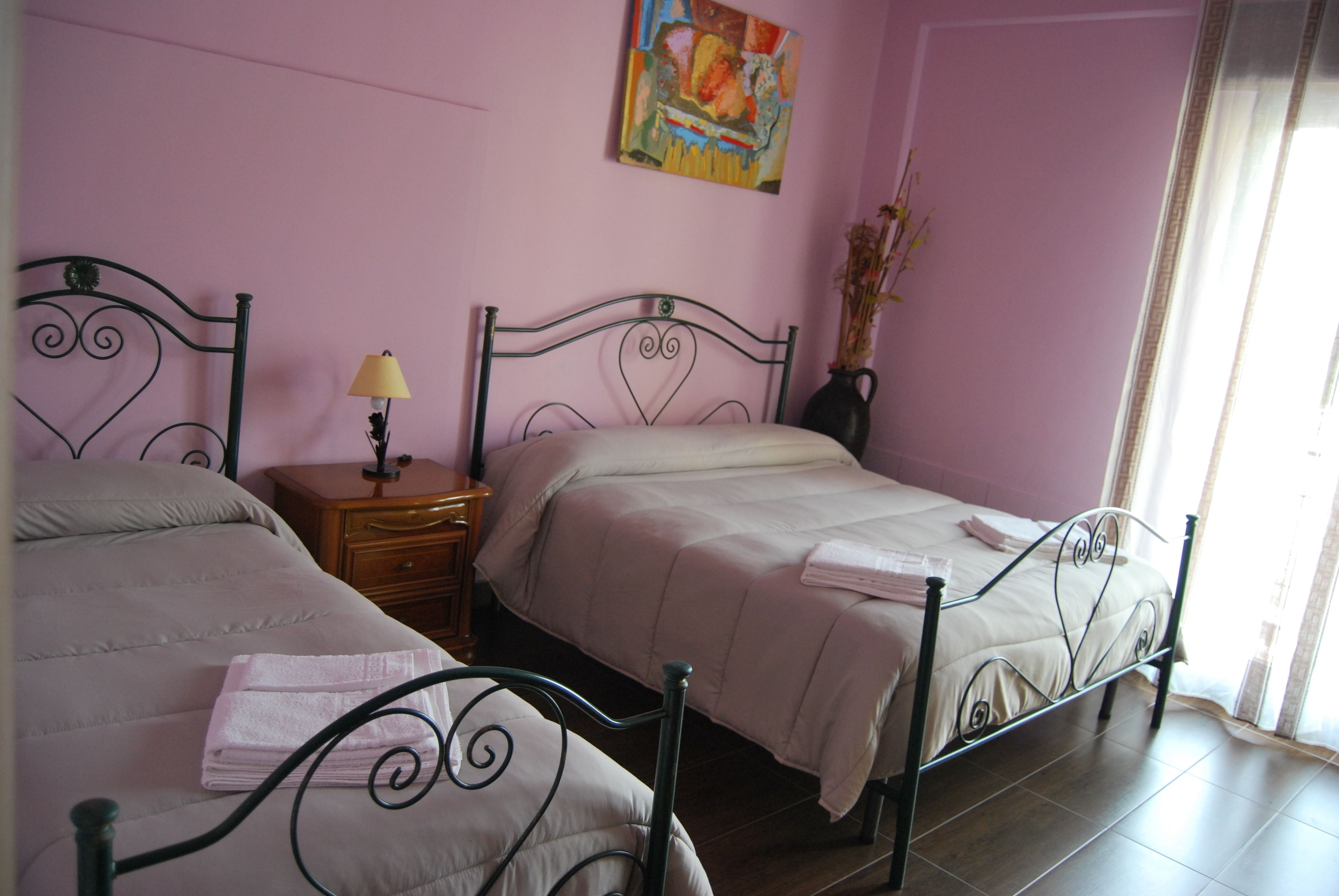 A bedroom with two beds, a nightstand with a lamp, a vase with flowers, and a painting on the wall.
