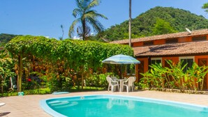 A swimming pool with lounge chairs and a table, surrounded by lush greenery and a building with a thatched roof. - Pousada Quatro Estações Paraty (Paraty)