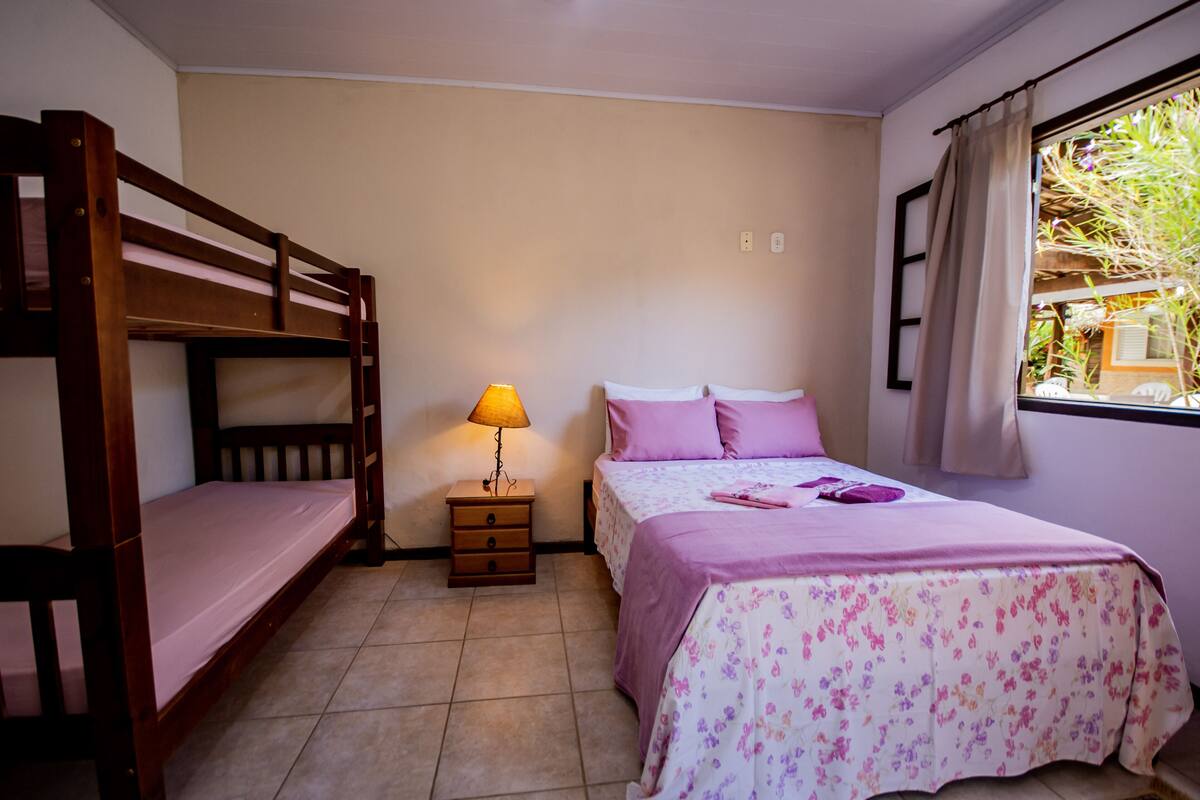 A bedroom with a bunk bed, a single bed, a nightstand with a lamp, and a window with curtains.