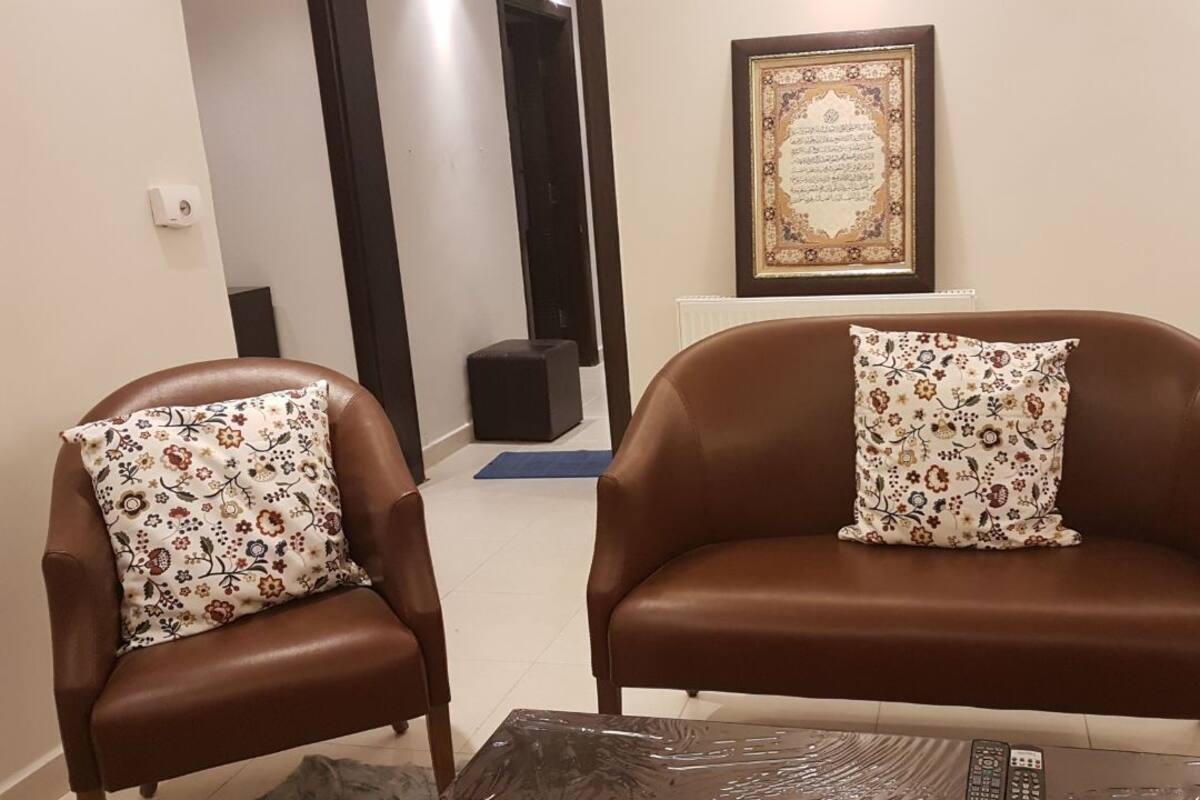 Ein Wohnzimmer mit zwei braunen Ledersofas, jedes mit einem floralen Kissen. An der Wand über den Sofas befindet sich ein gerahmter Koran.
