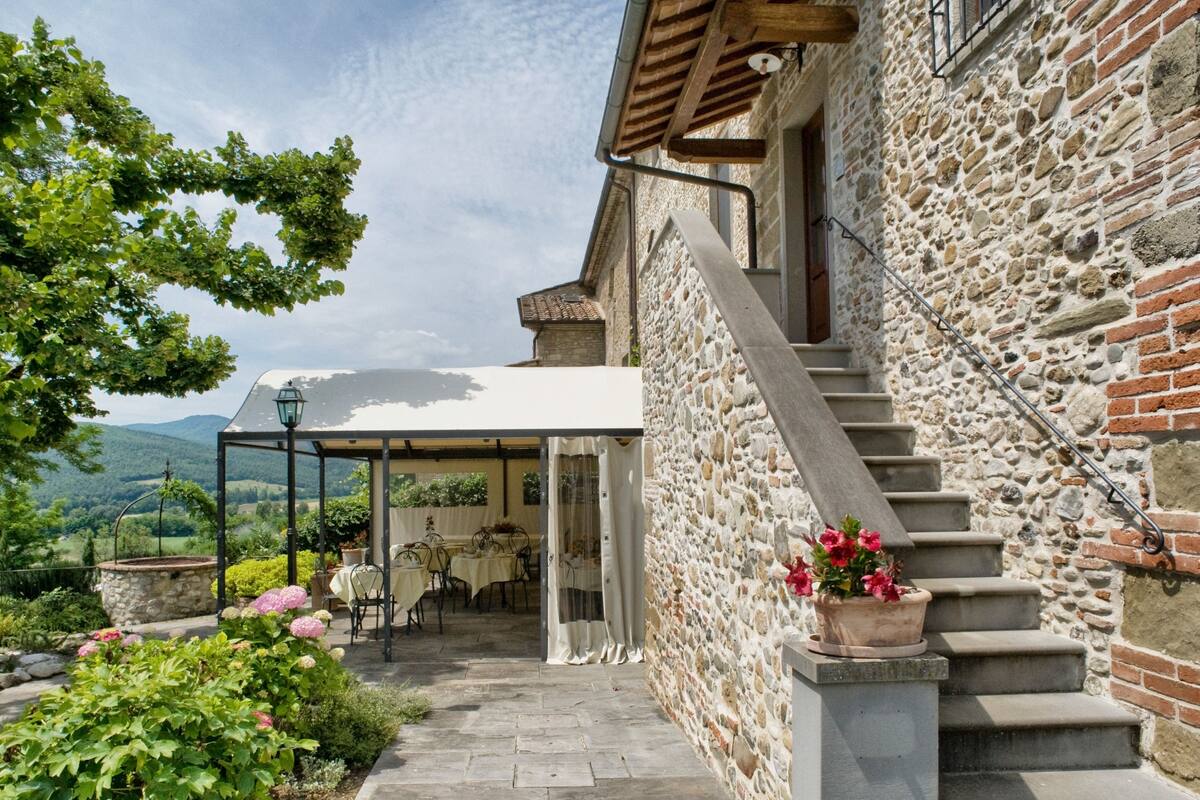 A stone building with a covered outdoor seating area, a staircase, and a view of the countryside.