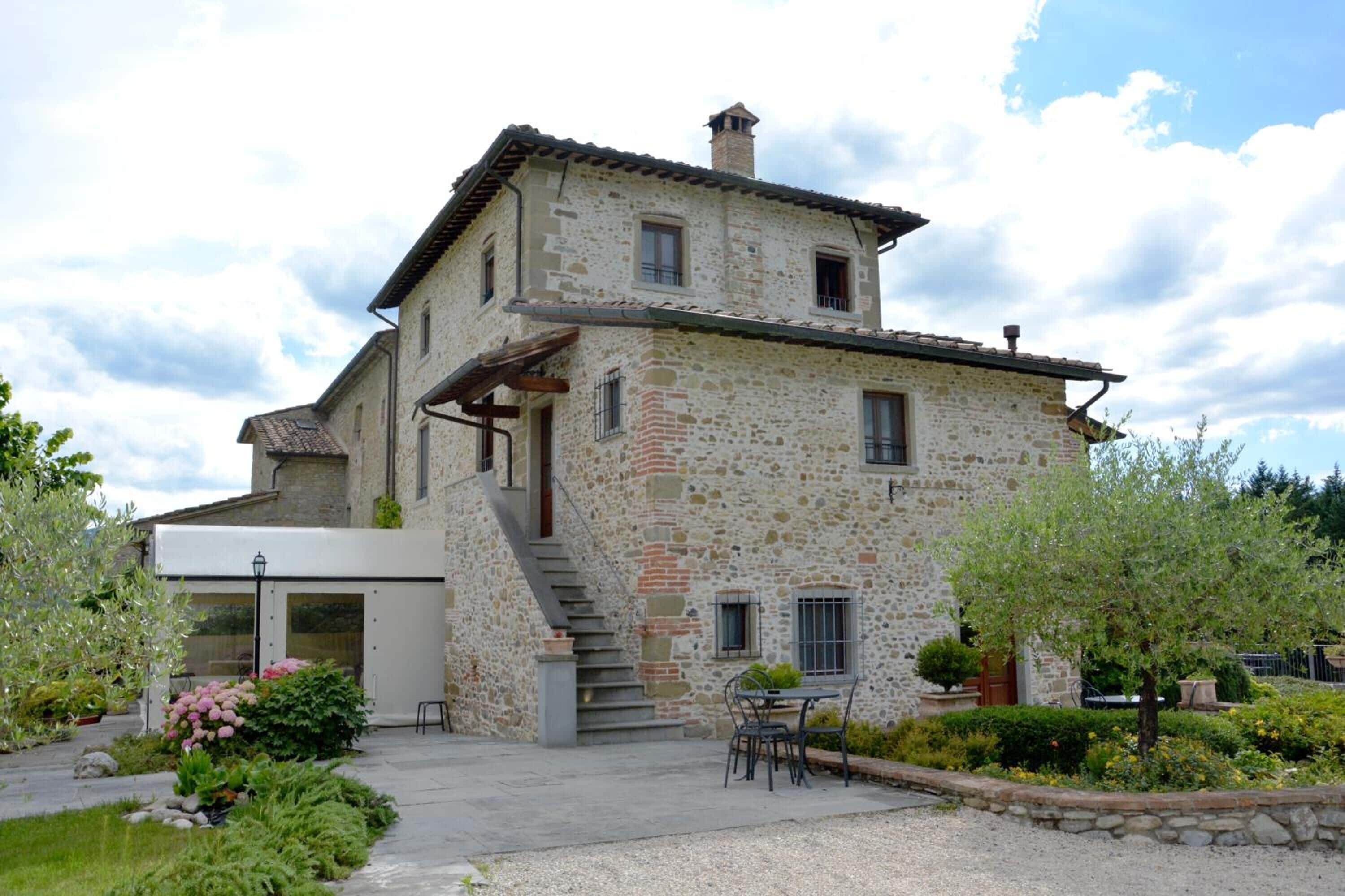 A stone house with a staircase leading to the upper floor, a patio with a table and chairs, and a garden with various plants.