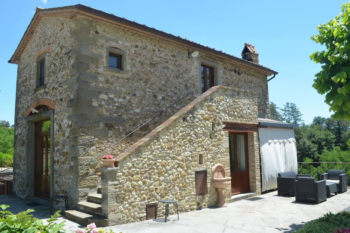 A stone building with a staircase leading to a door, outdoor seating, and a small garden area.