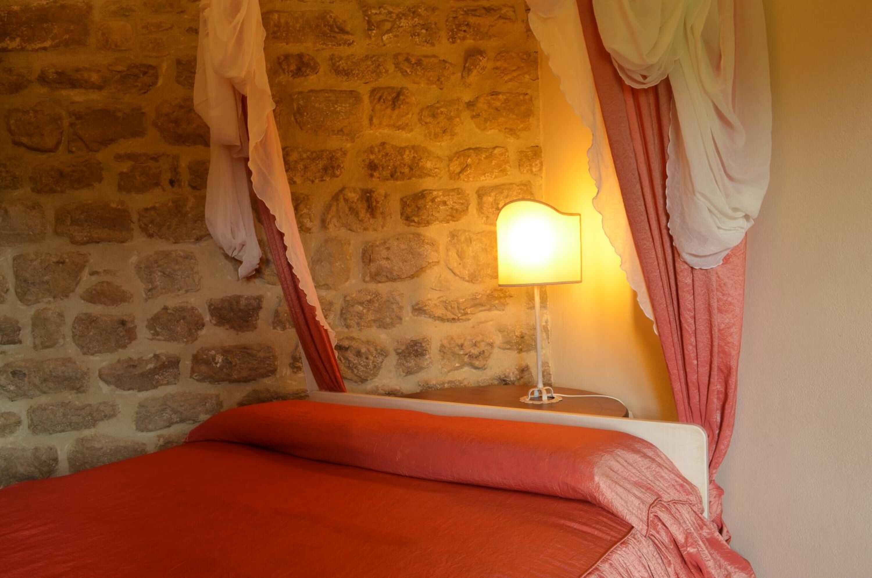 A room with a stone wall, a bed with a red blanket, and a bedside lamp.