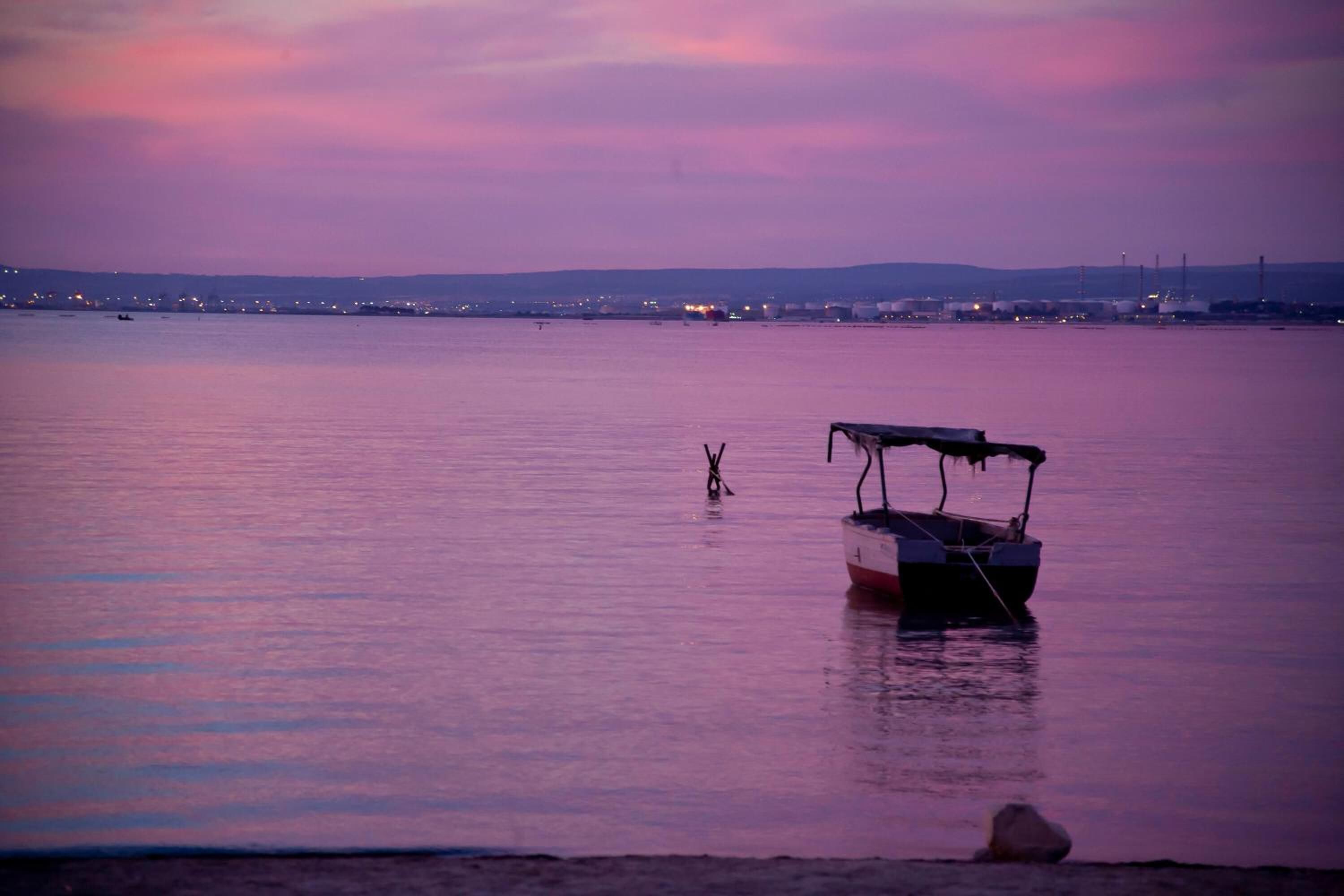 Ein Boot liegt in ruhigem Wasser vor einer Küstenlinie mit entfernten Industriegebäuden.