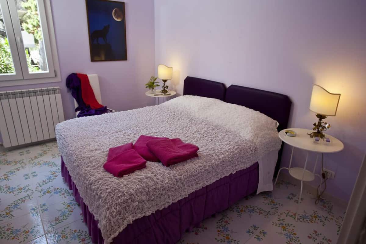 A bedroom with a large bed, two bedside tables with lamps, a radiator, a window with a view of greenery, and a painting on the wall.