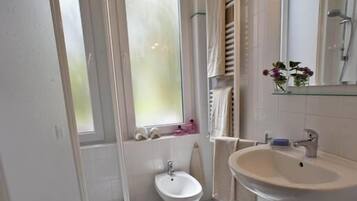Un baño con inodoro, bidé y lavamanos. Hay una ventana con vidrio esmerilado, una zona de ducha y un espejo sobre el lavamanos.