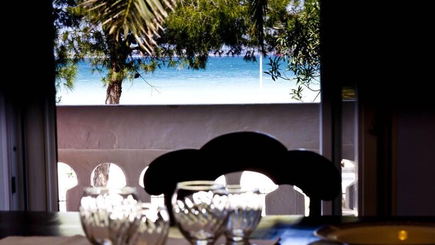 A dining table with glassware and a view of a beach and palm tree through a window. - La Villa della Meda (Taranto)