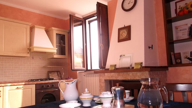 A kitchen with a stove, oven, and sink. There is a window with curtains, a clock on the wall, and a shelf with books and decorative items. - Dolcedorme B&B (Terranova di Pollino)