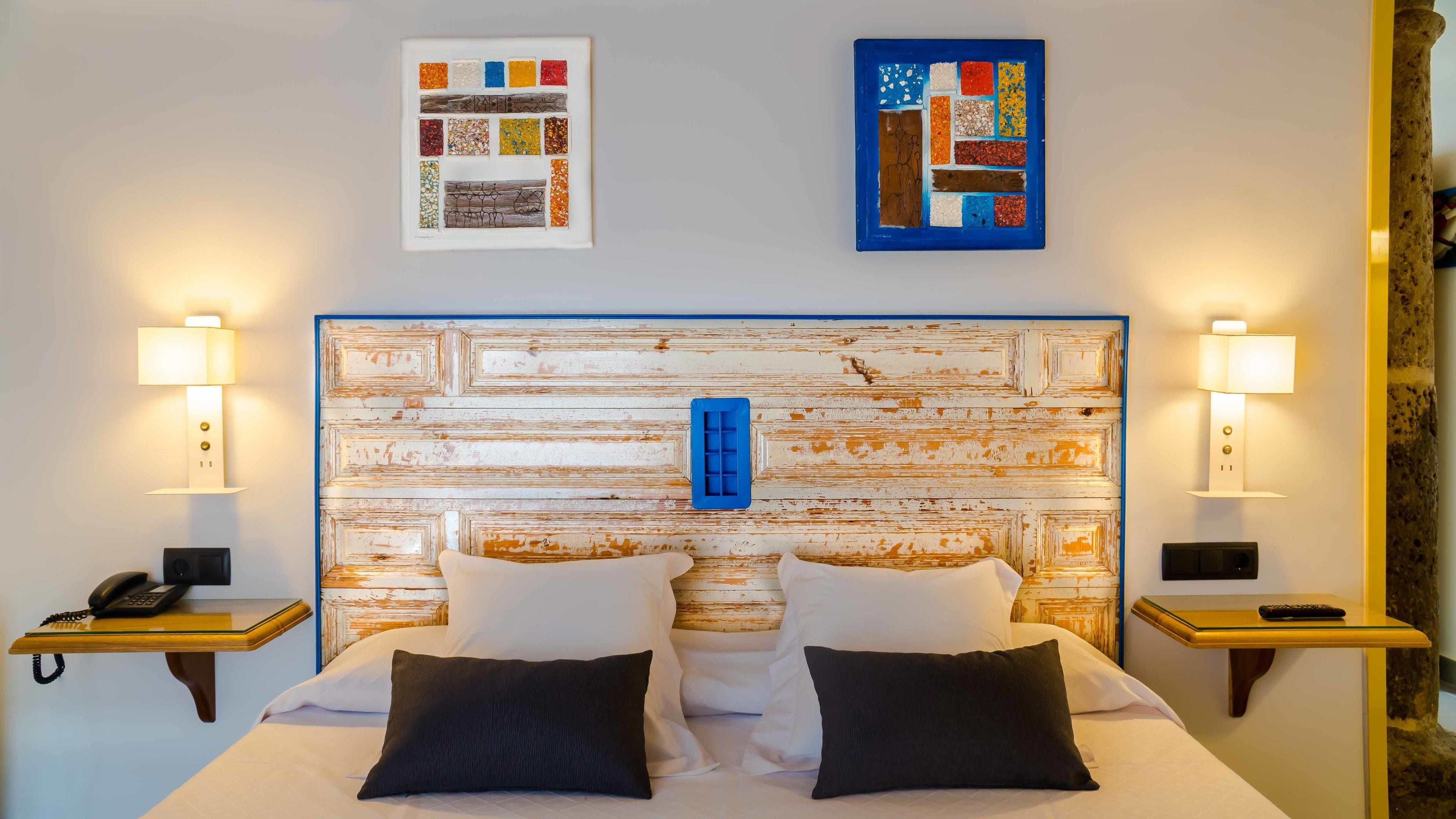 Una habitación de hotel con un cabecero de madera rústica, dos mesitas de noche con lámparas, una cama con sábanas blancas y almohadas negras, y obras de arte en las paredes.