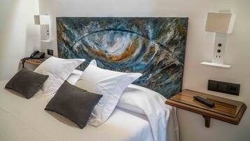 A hotel room with a large abstract painting on the headboard, a bed with white and gray bedding, two bedside tables with lamps and a remote control, and a wall socket.