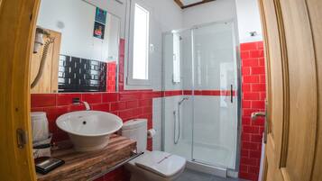 Un baño con un lavamanos blanco, paredes con azulejos rojos, un box de ducha de vidrio y una puerta de madera.