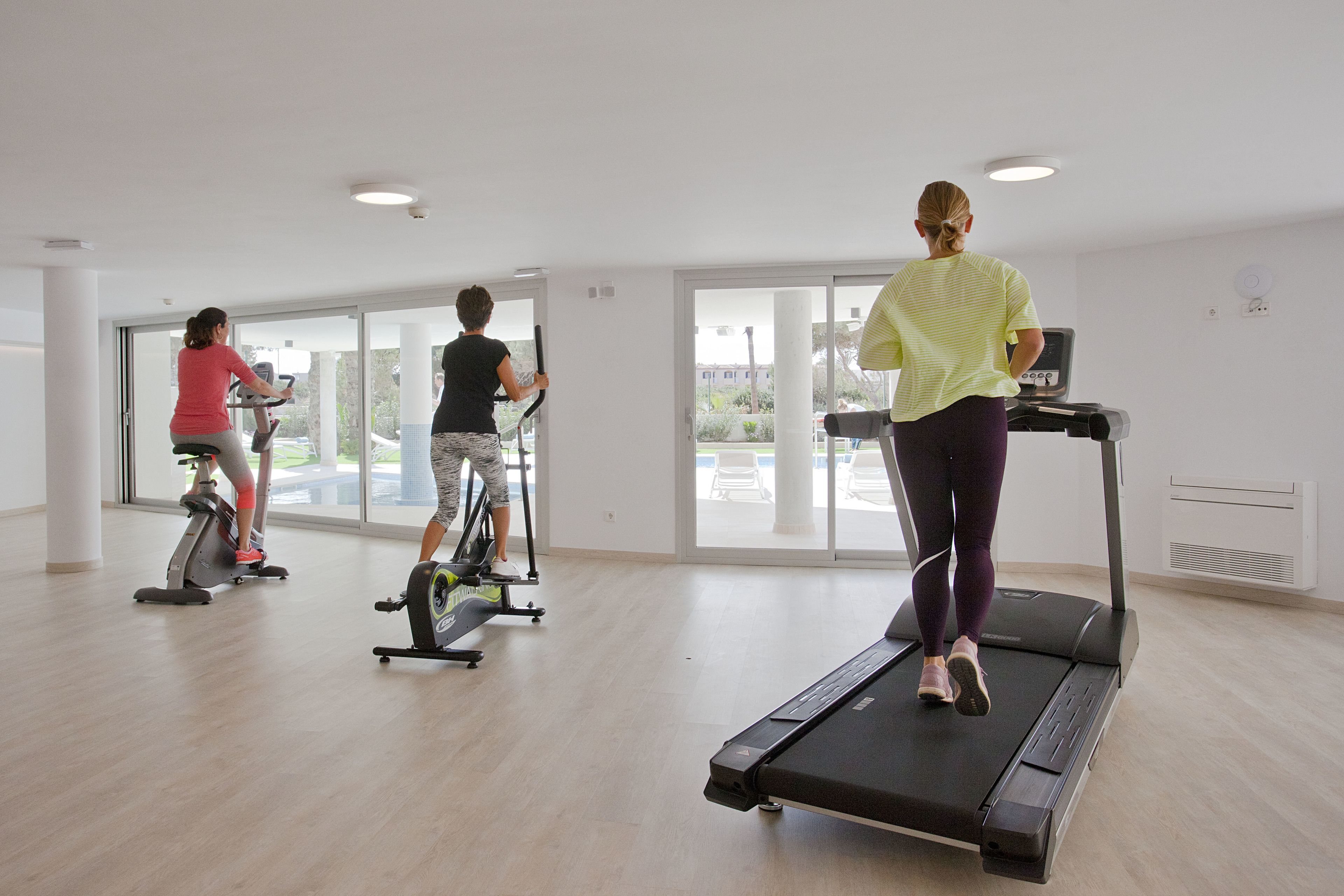 A gym with three people using cardio equipment, large windows, and a clean, modern interior.