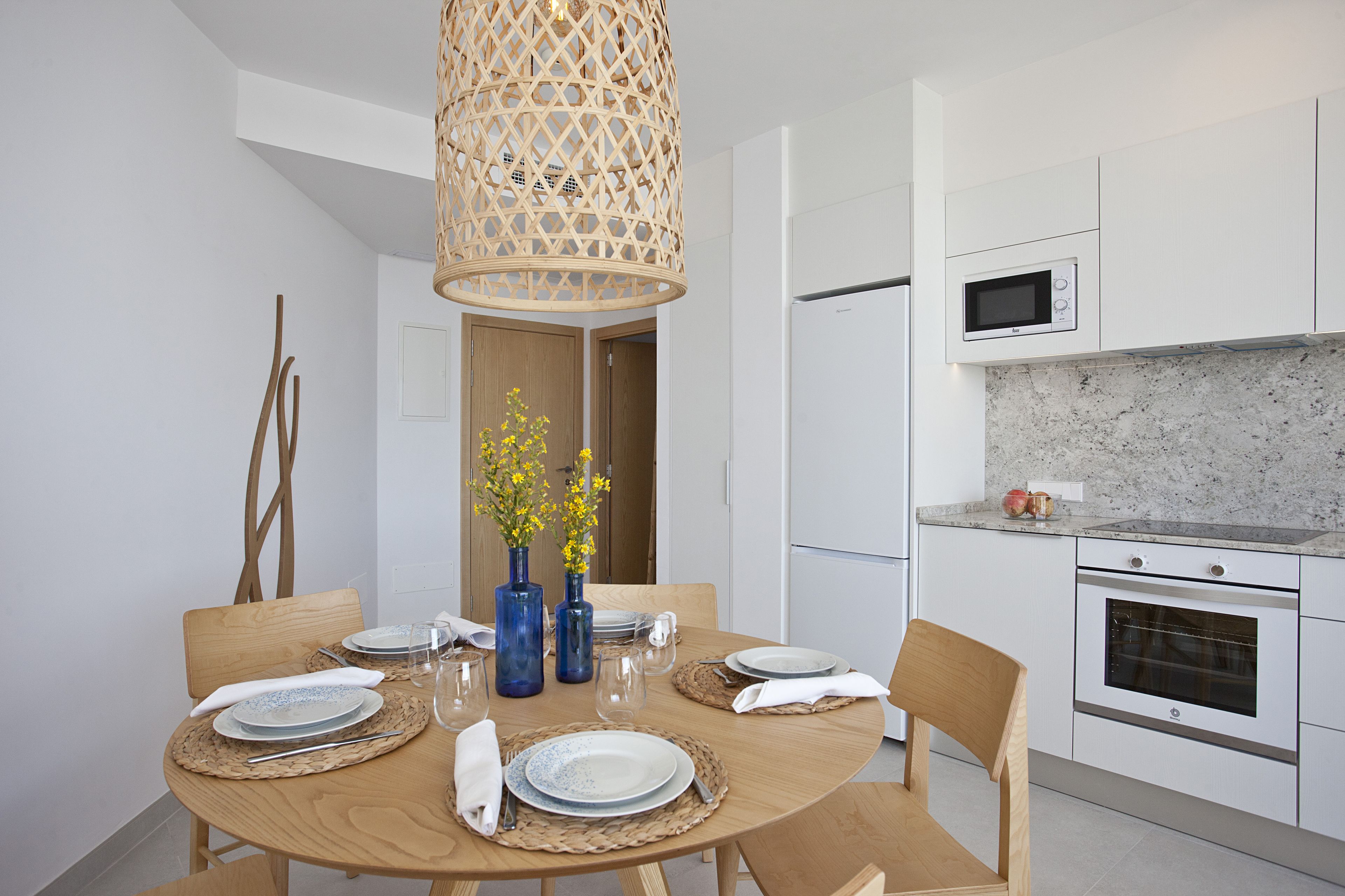 A modern kitchen with white cabinets, a microwave, and an oven. There is a dining table set with plates, glasses, and a vase with yellow flowers.