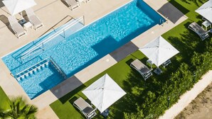 Vista aérea de un hotel con una amplia piscina rectangular, varias zonas de descanso con sombrillas y un césped bien cuidado.