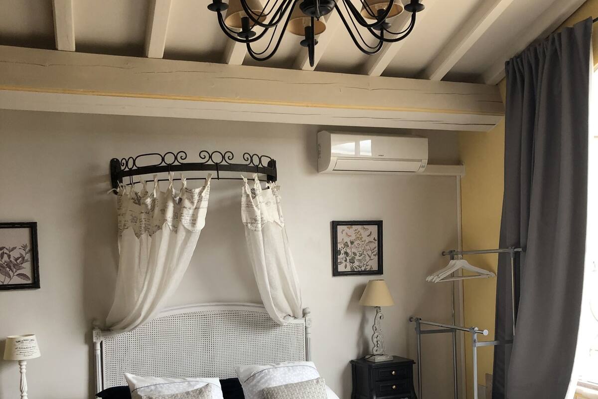 A bedroom with a four-poster bed, a standing lamp, a bedside table, and a window with curtains.