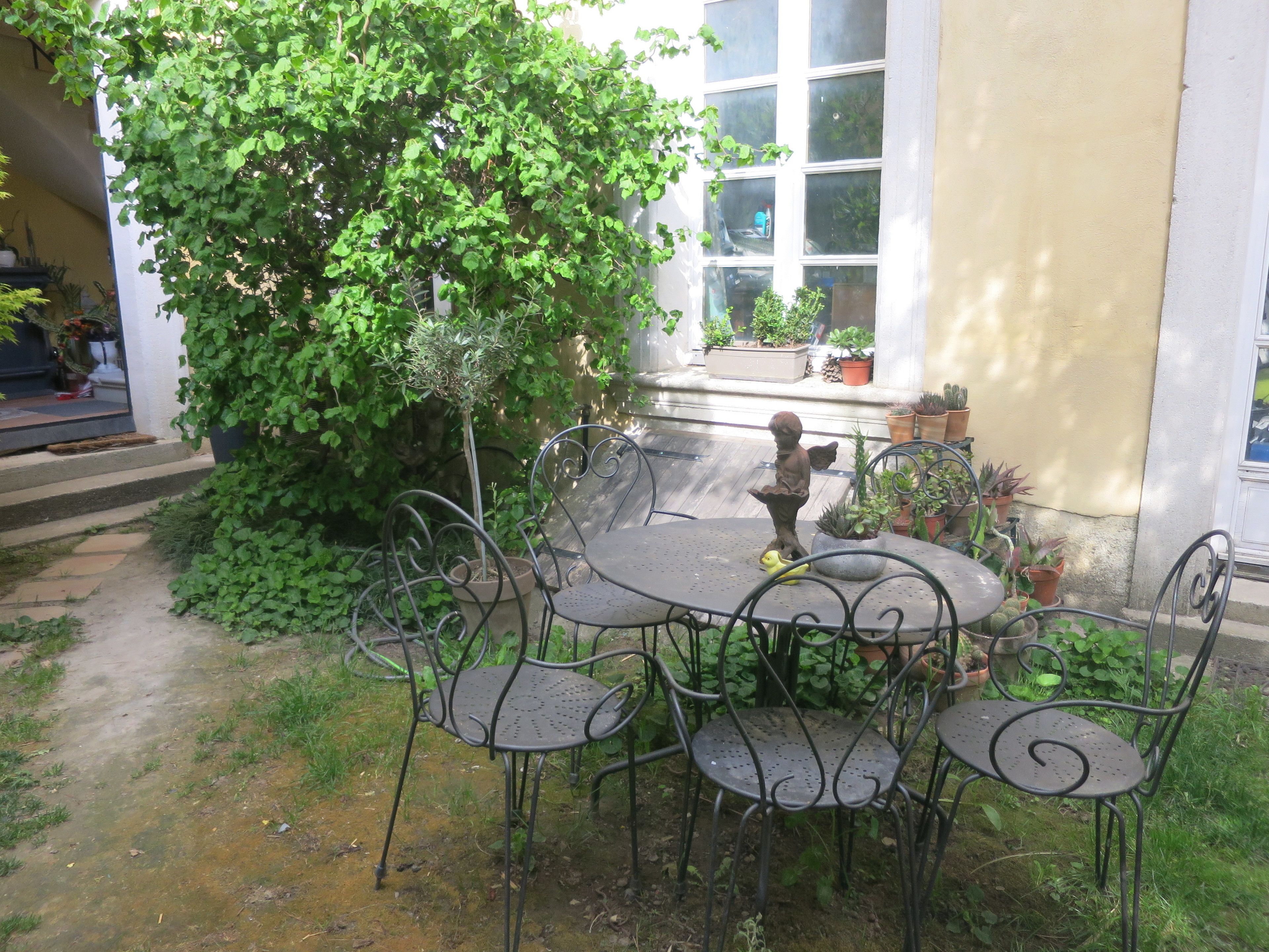 En udendørs terrasse med et rundt bord og metalstole, omgivet af krukker med planter og en stor busk.