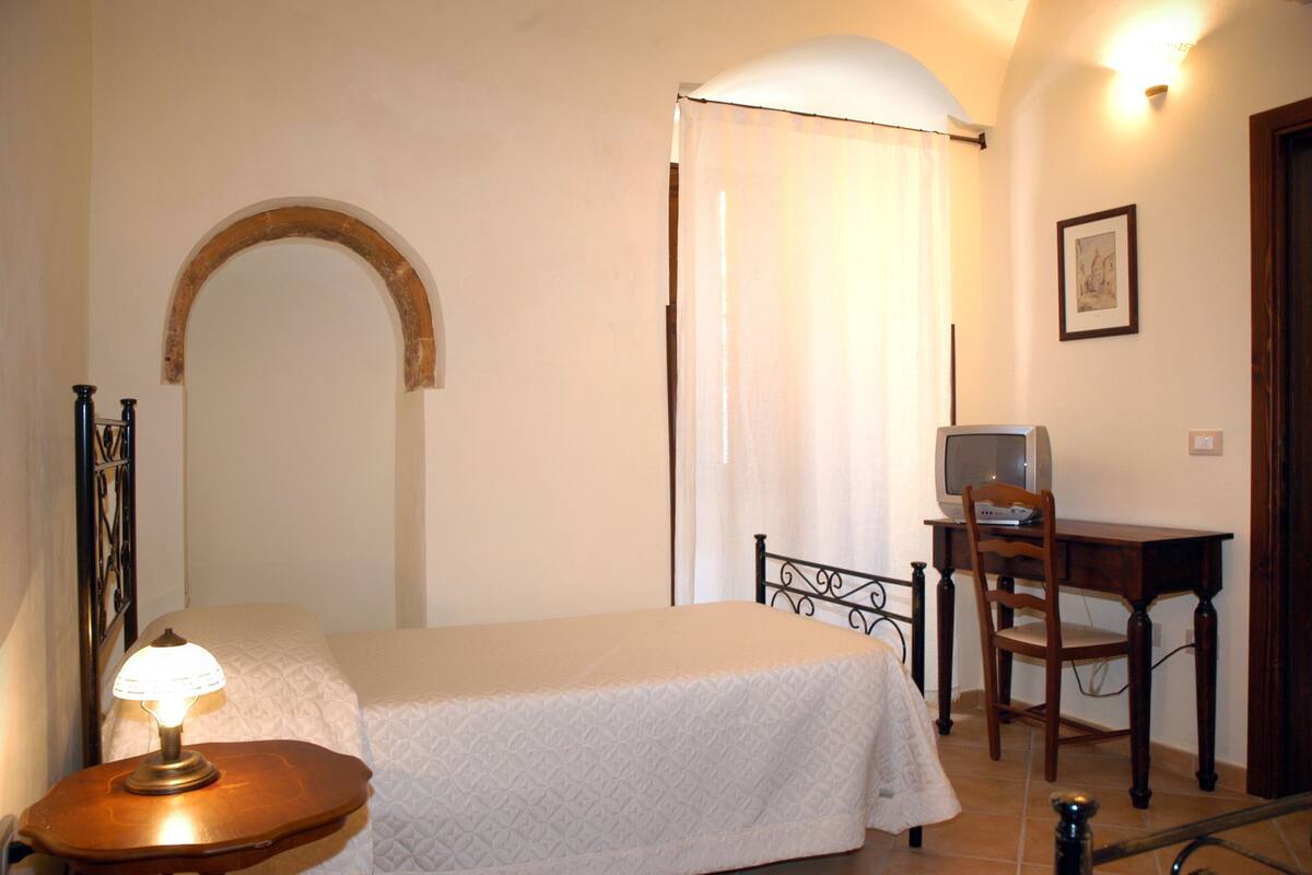 A bedroom with a bed, a desk with a chair, a lamp, and a picture on the wall.