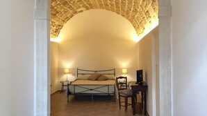 A bedroom with a stone archway, a bed, bedside lamps, a desk with a chair, and a wicker chair. - B&B Macrina (Noto)