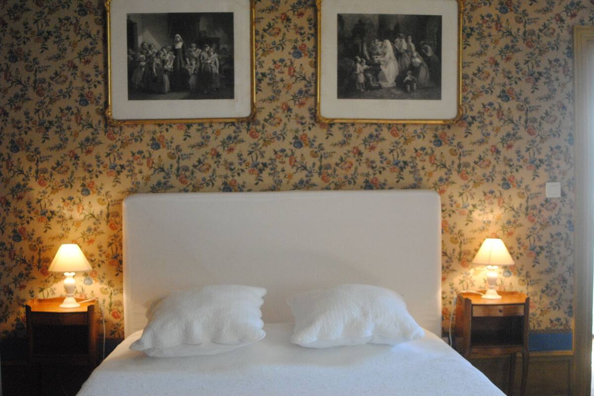 A neatly made bed with two pillows, flanked by two bedside tables with lamps, in a room with floral wallpaper and framed pictures on the wall.