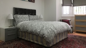 A bedroom with a bed, two bedside tables, a window, a painting, and a red patterned rug.