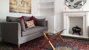 A living room with a grey sofa, red and patterned cushions, a glass coffee table, a large world map painting, a mirrored octagonal clock, and a white fireplace with a black birdcage.
