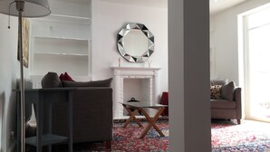 A living room with a grey sofa, a glass coffee table, a patterned rug, a white fireplace, and a round mirror on the wall.