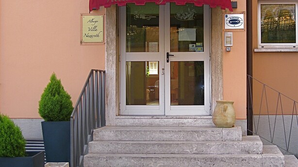 Entrada de un edificio con toldo rojo, un letrero que dice "Villa Nazareth" y una pequeña planta en maceta junto a las escaleras.