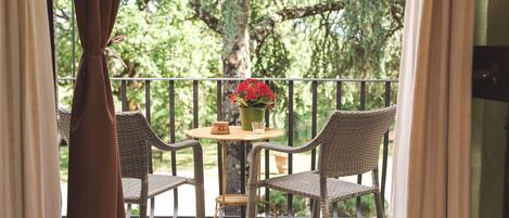 A balcony with wicker chairs, a small table with a vase of red flowers, and a view of trees.