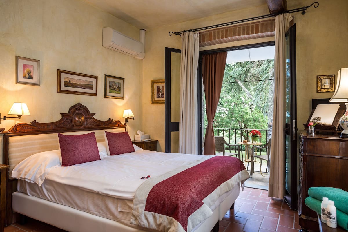 A hotel room with a large bed, a balcony with a view of trees, and a desk with a lamp.
