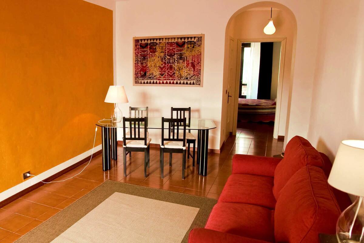 A living room with a red sofa, a dining table with two chairs, a lamp, and a colorful abstract painting on the wall.