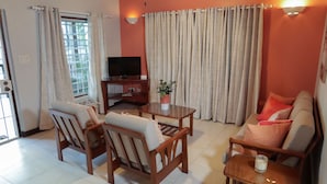 A living room with a sofa, two armchairs, a coffee table, a TV on a stand, and a potted plant. - Trilogy Villas (Falmouth Harbour)