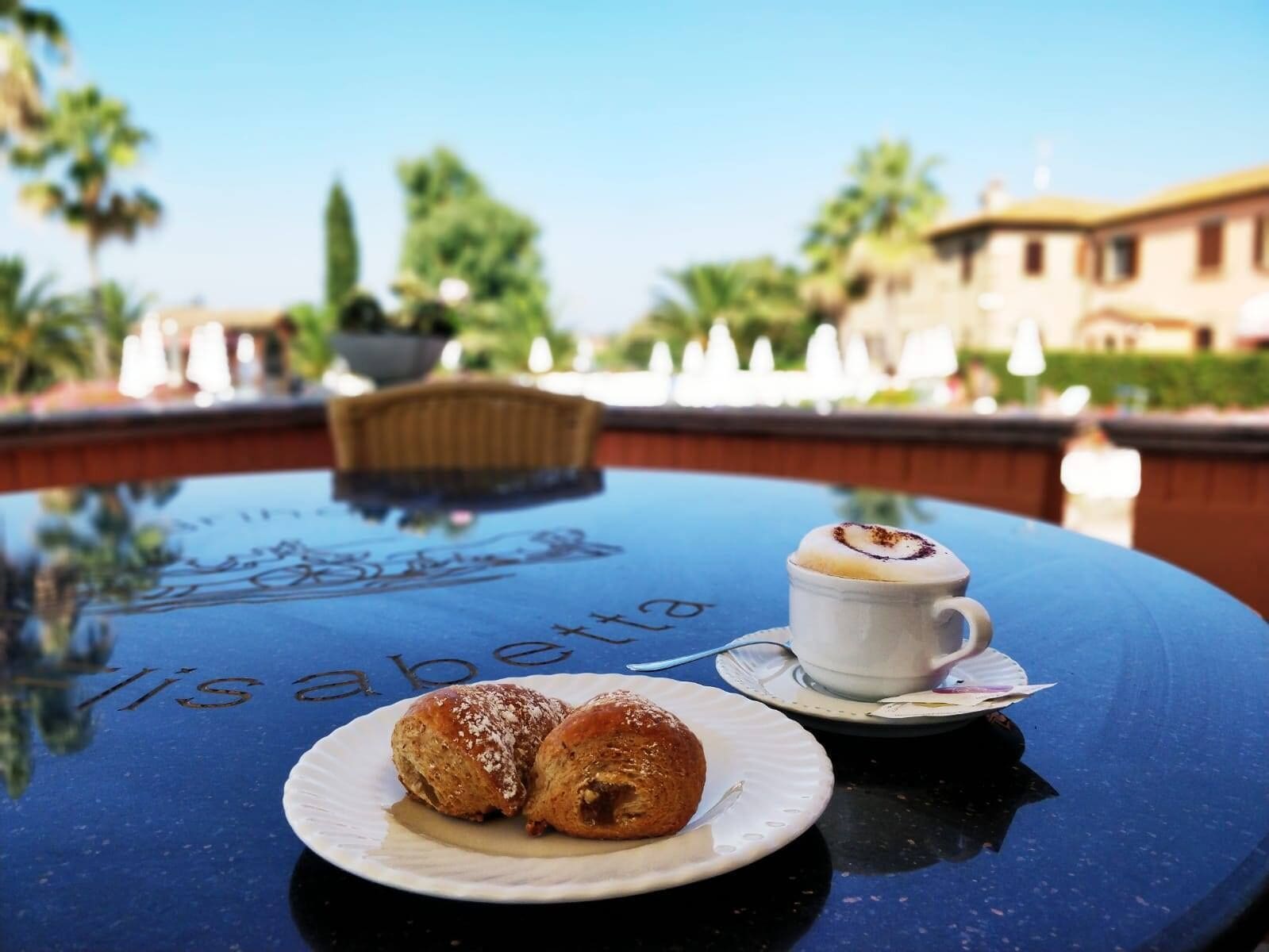Una mesa con un plato de pasteles y una taza de café, con una piscina y edificios al fondo.