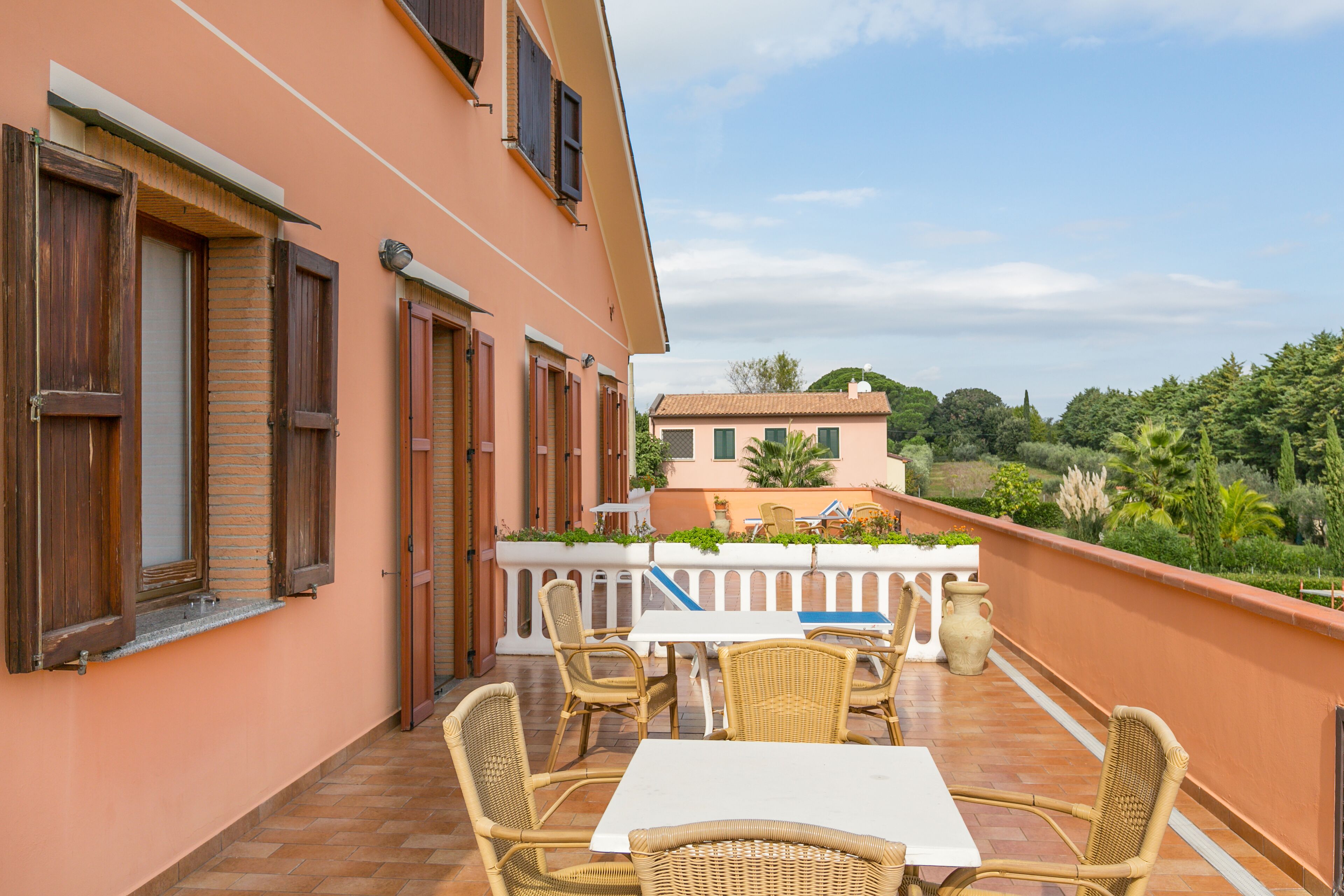 A balcony with wicker furniture and a white table, overlooking a garden and a building with a tiled roof.