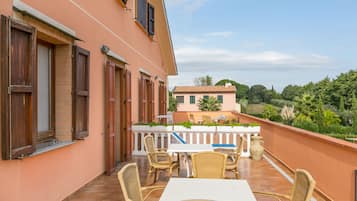 A balcony with wicker furniture and a white table, overlooking a garden and a building with a tiled roof.