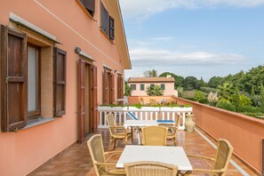 A balcony with wicker furniture and a white table, overlooking a garden and a building with a tiled roof. - Tenuta Elisabetta (Cecina)