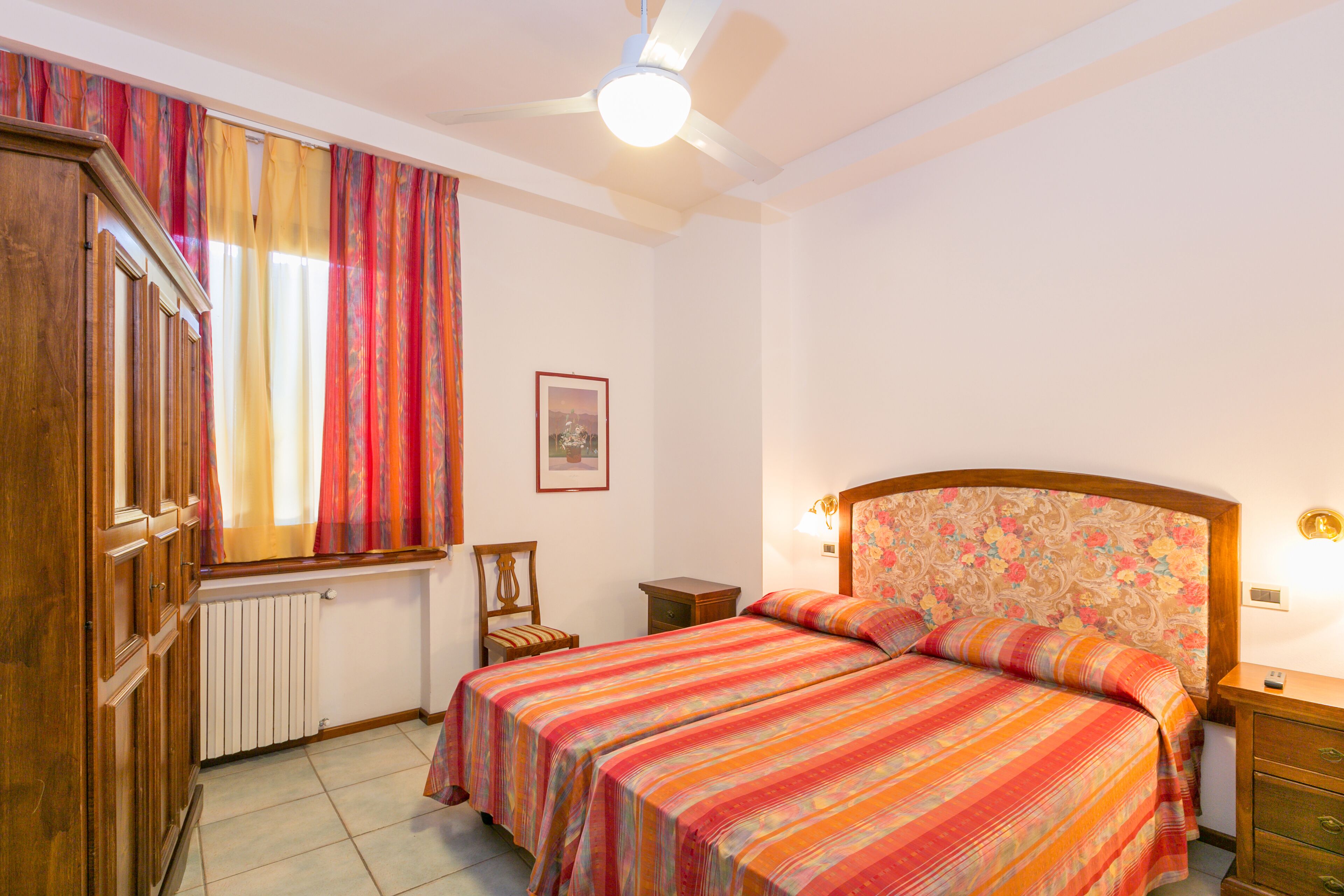 A bedroom with two beds, a wooden wardrobe, a radiator, a chair, and a bedside table with a lamp.