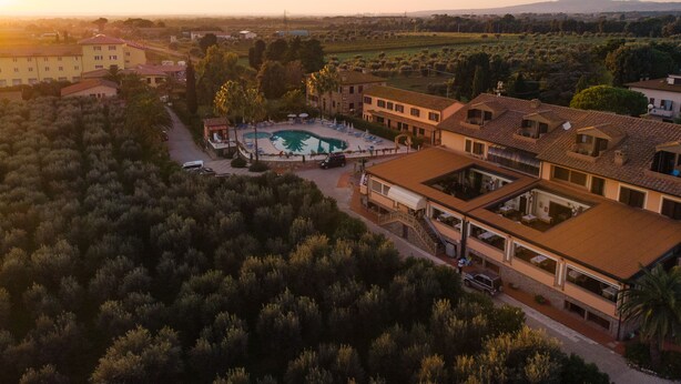 An aerial view of a resort with a swimming pool surrounded by buildings, a parking area, and a hilly landscape in the background. - Tenuta Elisabetta (Cecina)