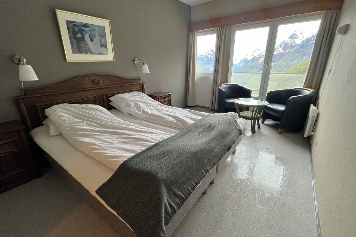 A hotel room with a large bed, a small table, two chairs, a lamp, and a painting on the wall.