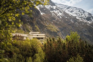 Un edificio moderno con tetto piatto, immerso in una fitta foresta e affiancato da una montagna con chiazze di neve.