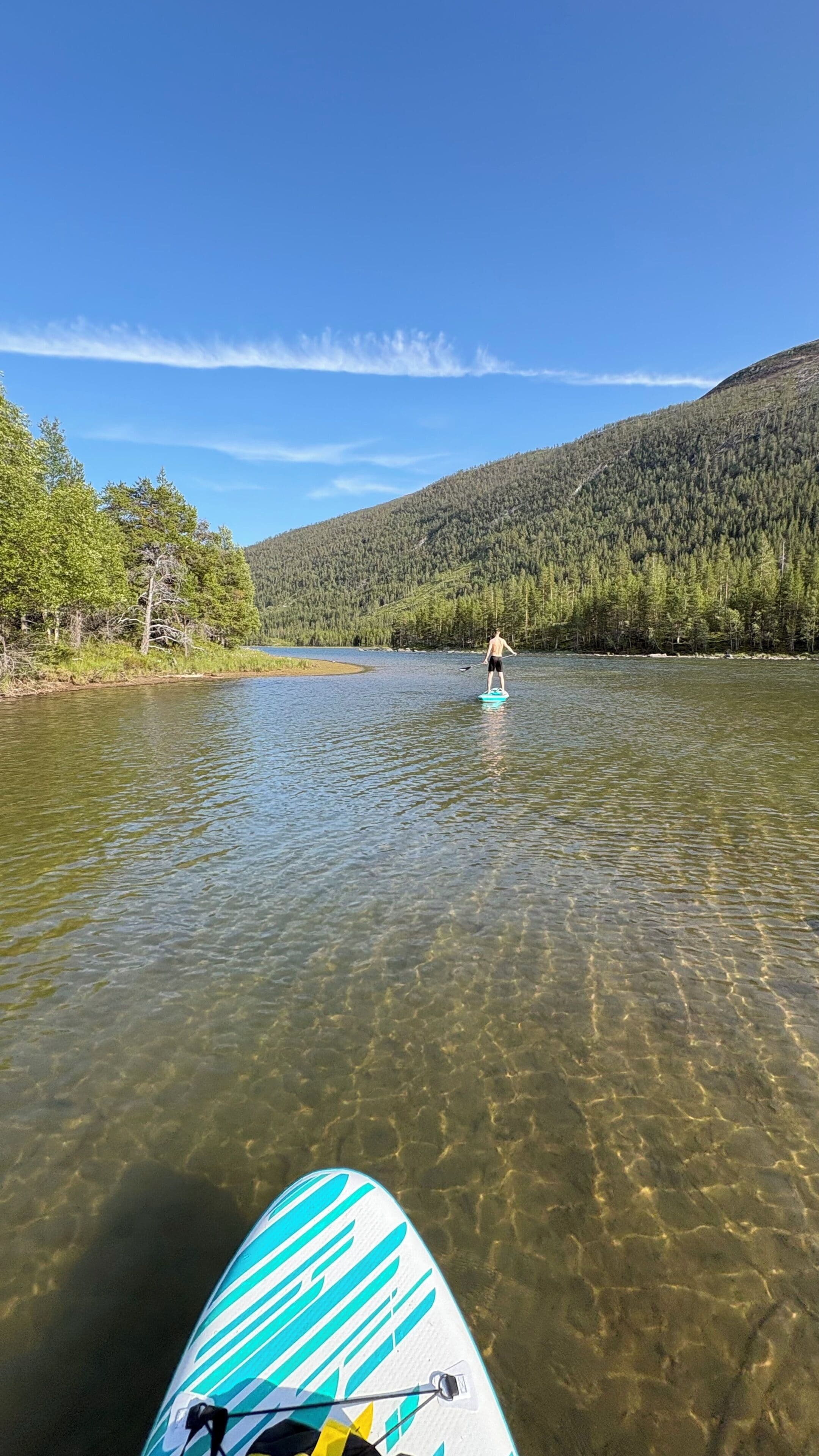 A person paddleboarding on a calm river with clear water, surrounded by forested hills.