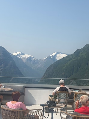 Une terrasse dotée de chaises et de tables en osier, offrant une vue sur un paysage montagneux aux sommets enneigés.