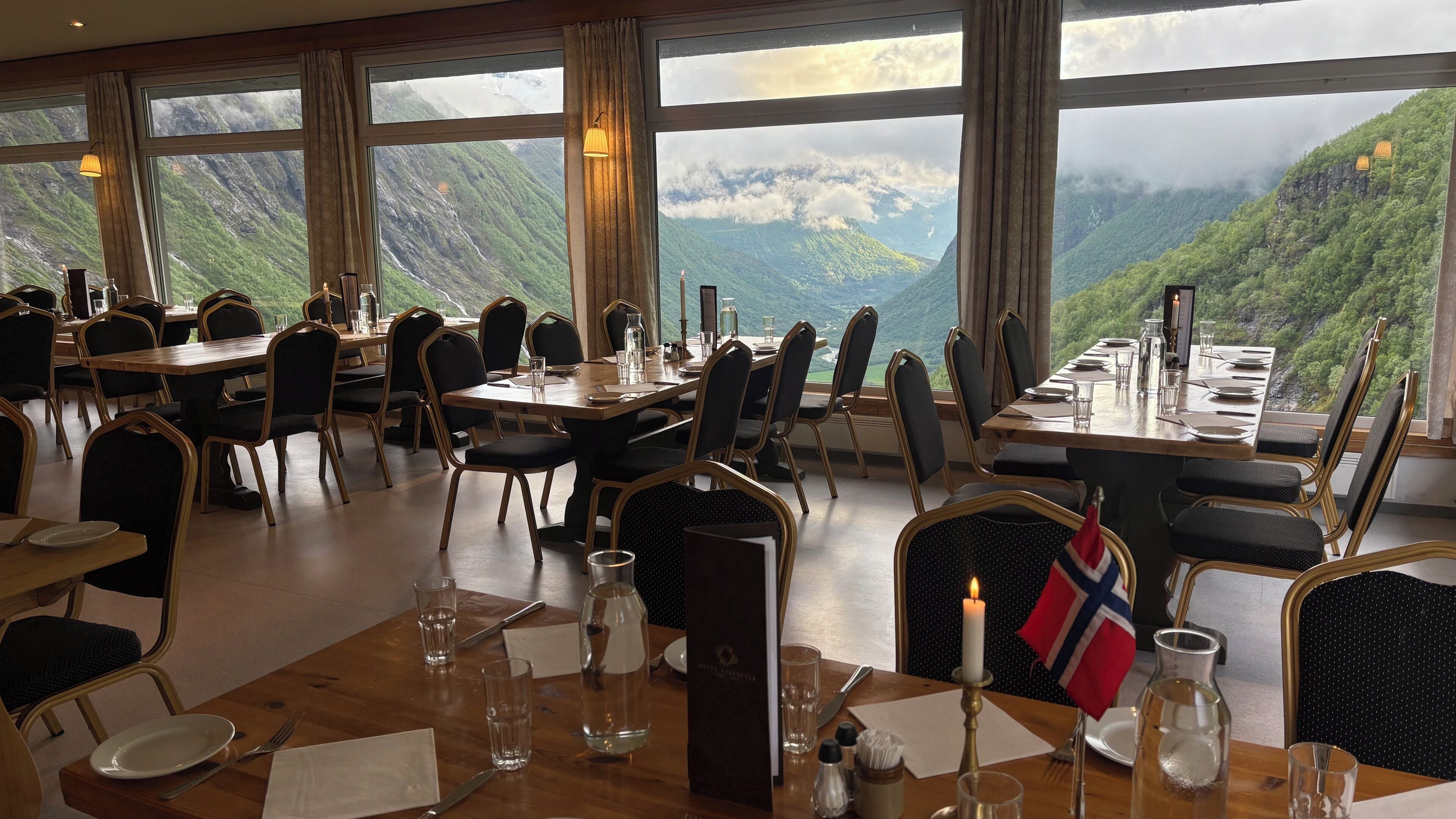 Un restaurante con mesas dispuestas para comer, amplias ventanas que ofrecen vistas a la montaña y una bandera sobre una mesa.