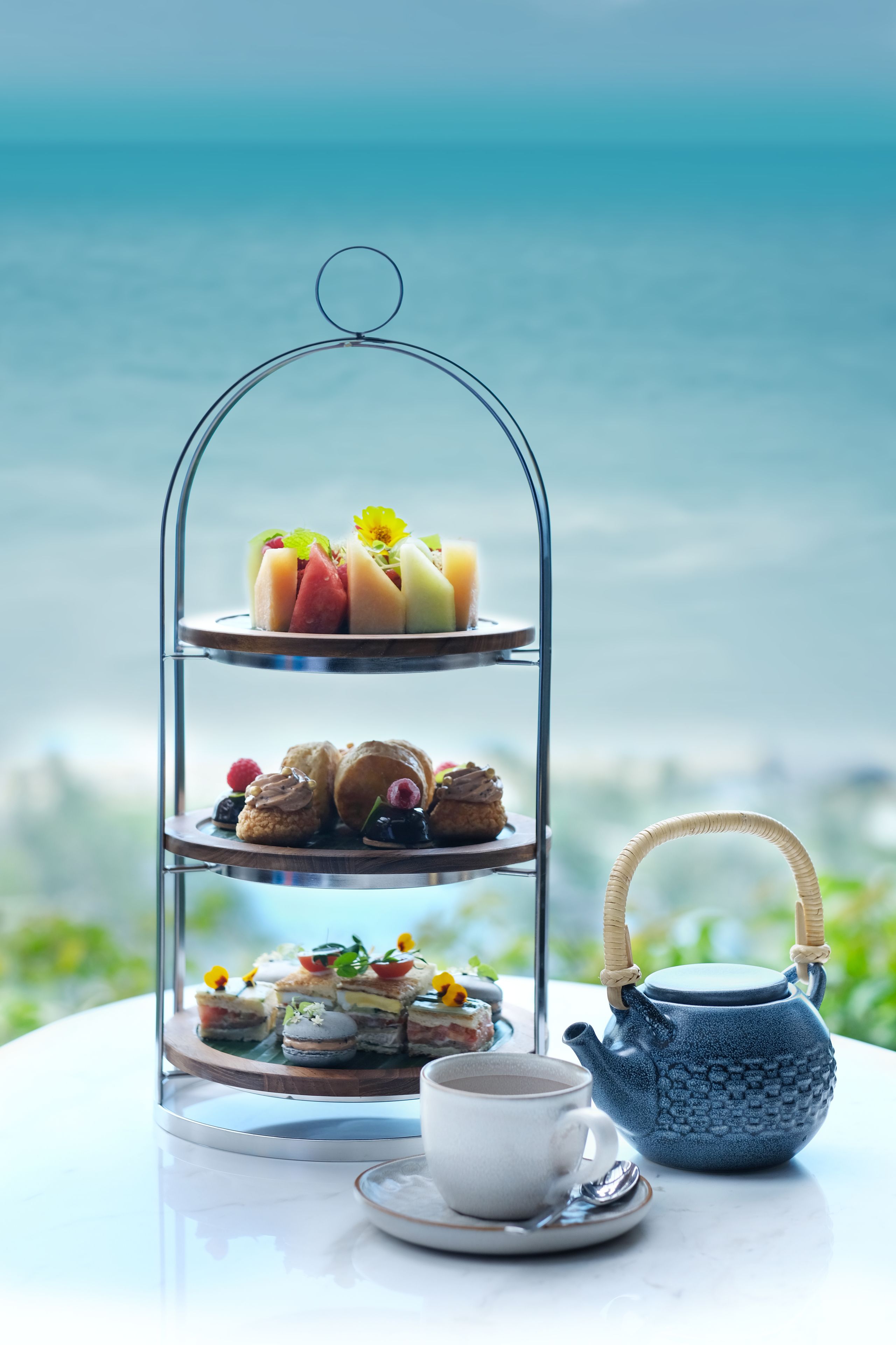 A three-tiered dessert stand with assorted pastries and a teapot on a table with a view of the ocean.