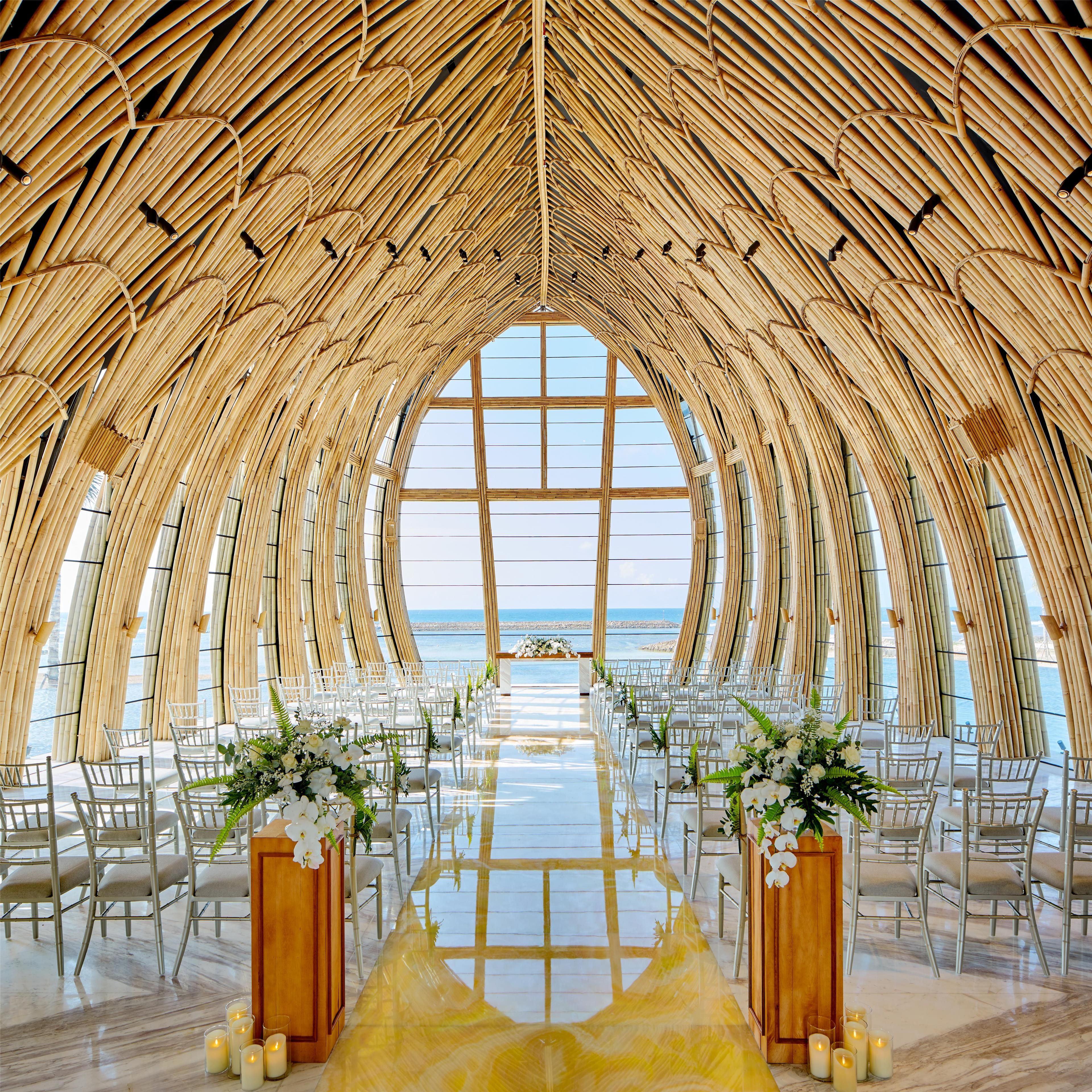 An indoor venue with a high ceiling made of bamboo, a central aisle with chairs, and a large window offering a view of the sea.