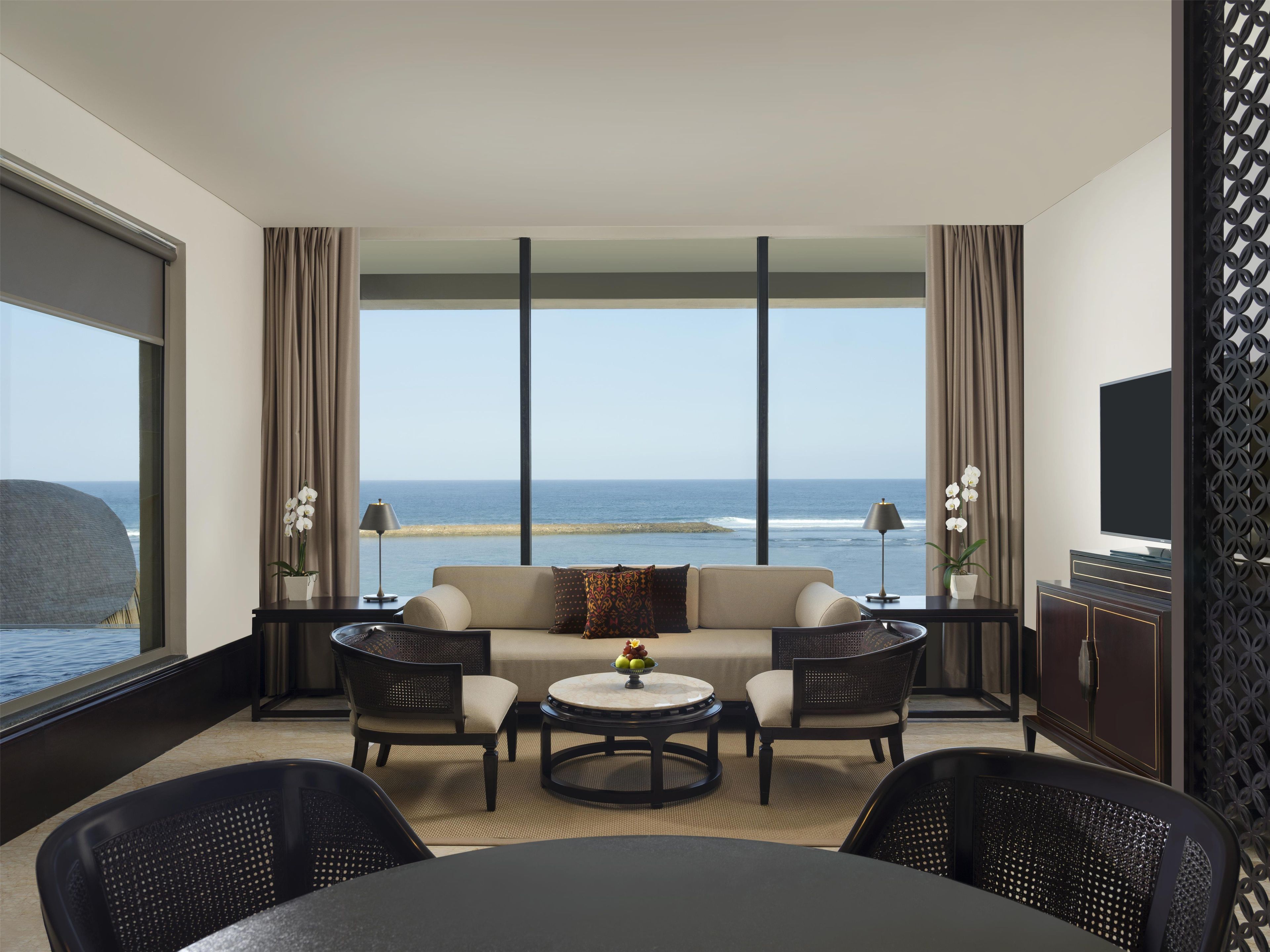 Habitación de hotel con sofá, dos sillones, una mesa redonda, televisor y vista al océano.